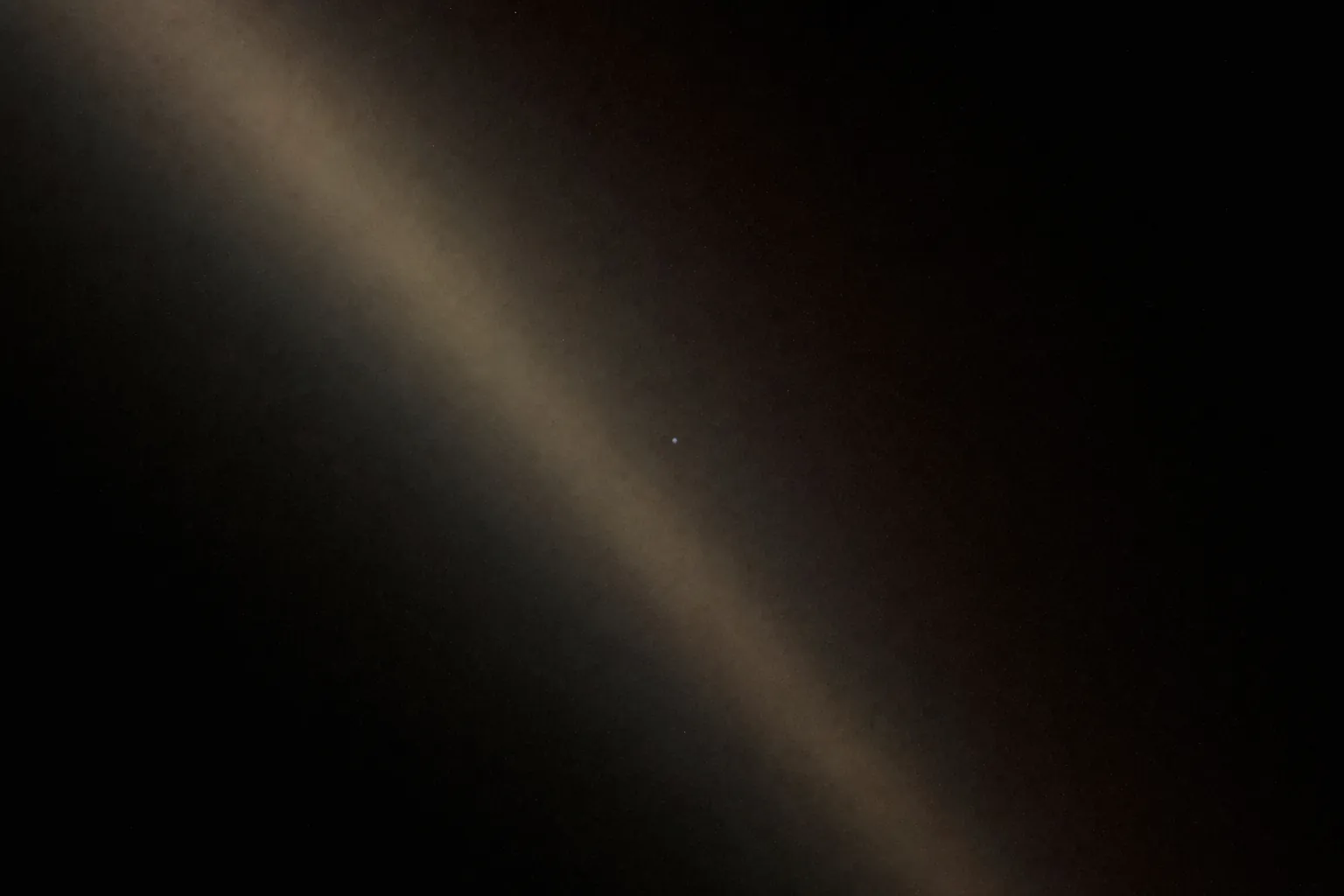 A faint pale blue point of light suspended in a broad diagonal band of scattered sunlight against black space.